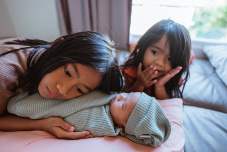 In a cozy setting, sisters watch over their adorable newborn sibling, creating sweet momentsの写真素材
