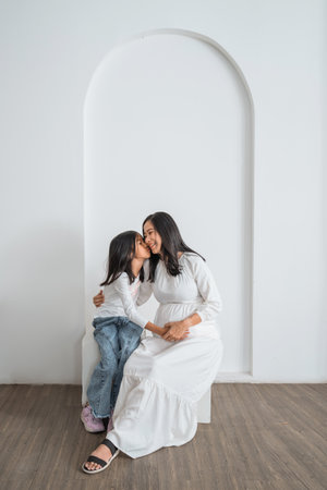 A pregnant woman is happily sitting alongside a little girl who is sweetly kissing her softly on the cheek, sharing a lovely moment togetherの写真素材