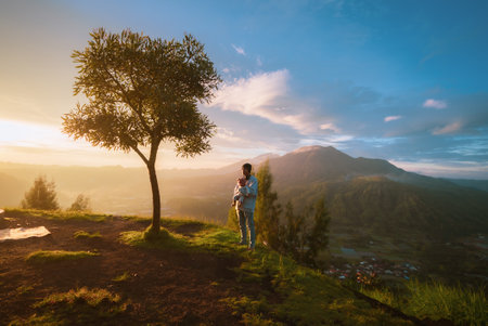A man is confidently standing atop a picturesque hill, next to a lush tree that complements the beautiful natural scenery around himの写真素材