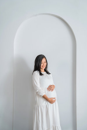 A beautiful pregnant woman wearing a flowing white dress is standing gracefully in front of a pristine white wall, gently holding her belly with both hands in a loving gestureの写真素材