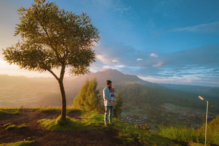 A man is standing on top of a high hill next to a solitary tree that is surrounded by a beautiful natural landscapeの写真素材