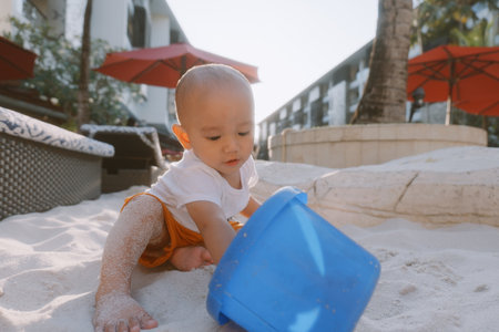 A happy baby is joyfully playing in warm sand with a blue bucket on a sunny dayの写真素材