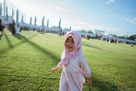 A cheerful young girl wearing a pink dress and a hijab plays joyfully in a sunny green fieldの写真素材