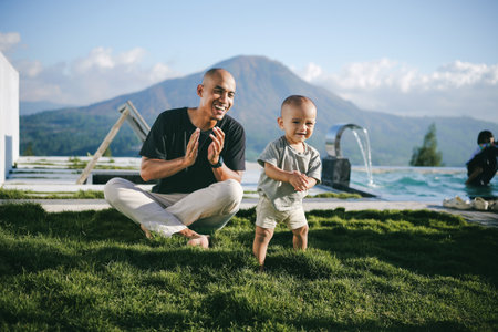 A man and a young child are comfortably seated on the lush green grass close to a sparkling swimming pool, enjoying their leisurely time togetherの写真素材