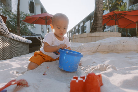 A child is enjoying playing in warm sand with a blue bucket and colorful toys aroundの写真素材