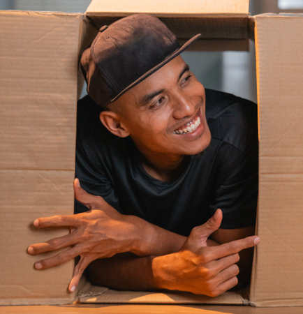 A cheerful young man smiles and poses playfully from inside a cardboard box, radiating joyの写真素材