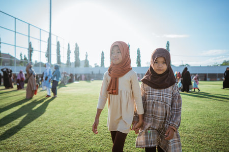 Two young girls in beautiful hijabs share joy in a sunlit gathering, surrounded by natureの写真素材