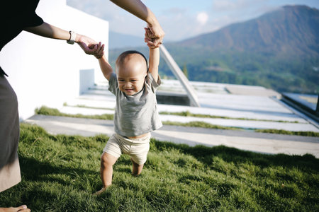 A woman gently holds the tiny hand of a baby while they both walk together on the soft grass,...