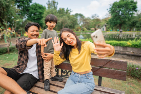 young mom takes cell phone selfie with father and little boy while sitting on park benchの写真素材