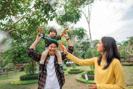 mom makes bubbles with a boy on his fathers shoulders while playing in the parkの写真素材