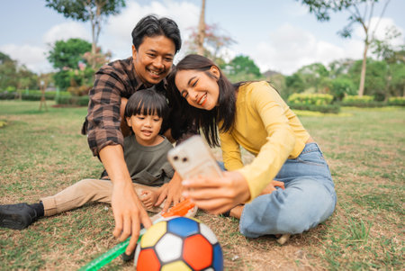 mother holding cell phone taking selfies of her son and father playing ball at the parkの写真素材
