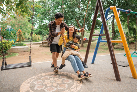 young woman holding little boy on her lap playing swing with young man in parkの写真素材