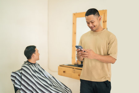A man is currently sitting in a barber shop getting his hair cut, while another nearby man is intently looking at his smartphone device in his handの写真素材