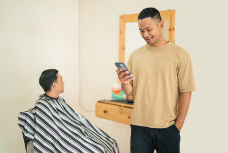 A man is standing closely next to another man who is seated in a barber chair, intently looking at his cell phone and checking messagesの写真素材