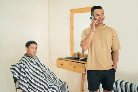 One man is engaged in a conversation on a cell phone, while another man is comfortably seated in a barber chair, preparing for a haircutの写真素材