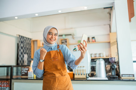 happy woman in headscarf with gesture gets surprise while looking at cell phone in coffee shopの写真素材