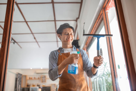 male employee wearing apron cleaning glass window with wiper and spray at coffee shopの写真素材
