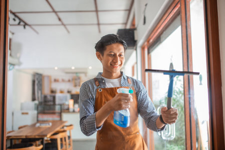 handsome male waiter in apron cleaning glass with wiper and spray in coffee shopの写真素材