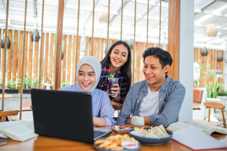 three Asian students laugh watching a laptop together in a coffee shopの写真素材