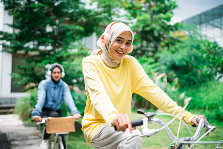 young woman in hijab smiling with headphones on bike while cycling with friends in parkの写真素材