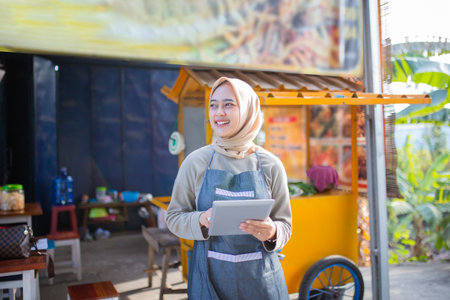 A smiling woman is happily using a tablet while standing at a vibrant, colorful food cartの写真素材