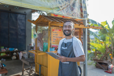 A joyful vendor is cheerfully taking orders at his vibrant food stall in a busy outdoor areaの写真素材