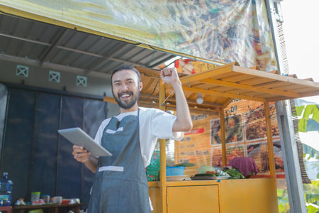 A joyful vendor proudly using a tablet at a vibrant street food stall, celebrating their successの写真素材