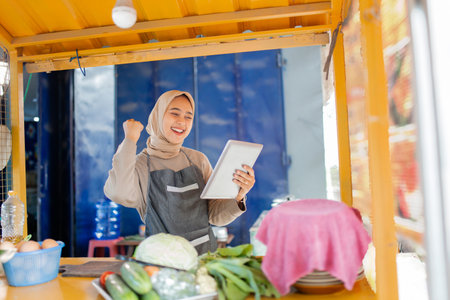 A joyful vendor celebrates at her vibrant food stall, using a tablet and attracting manyの写真素材