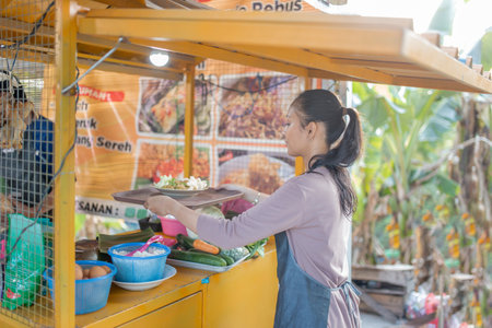 A woman at a bustling food stall is enthusiastically serving a variety of delicious dishesの写真素材