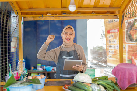 A joyful vendor showcases fresh produce and engages with customers while holding a tabletの写真素材