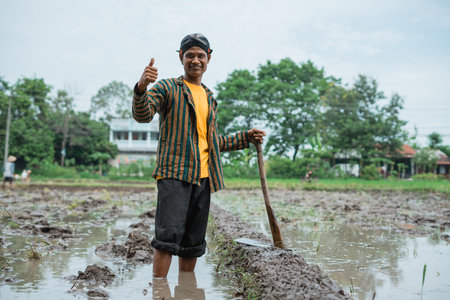 A joyful farmer stands in a picturesque wet rice field, joyfully signaling abundant positivityの写真素材