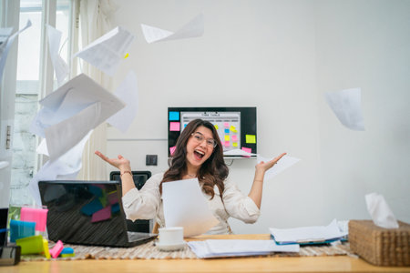 A cheerful woman is enjoying her success while surrounded by a flurry of paperwork and filesの写真素材
