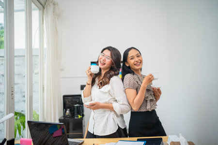 In a modern office, two cheerful women enjoy coffee together, fostering camaraderieの写真素材