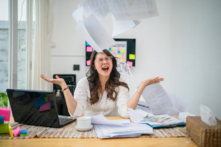 A stressed woman in a cluttered home office is throwing papers into chaos all around herの写真素材