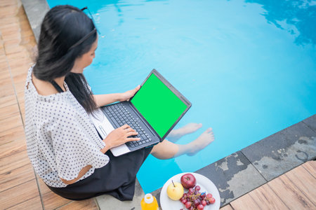 A woman enjoys working poolside, snacking and using her laptop, enhancing her relaxationの写真素材