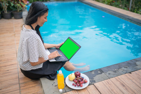 A woman enjoys a laptop work session by the pool, indulging in snacks and lemonadeの写真素材