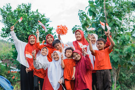 excited young girl in hijab and smiling boy holding small Indonesian flag during independence...