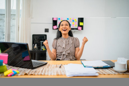 A businesswoman joyfully celebrates her achievement in her stylish, modern workspaceの写真素材