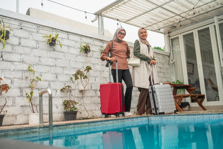 Two women standing with luggage beside a beautiful pool in a stylish outdoor area.の写真素材