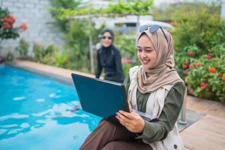 A woman is happily using her laptop by the poolside, enjoying the beauty of nature surrounding herの写真素材