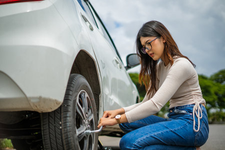 A woman is actively engaged in repairing the tire of her car in a sunny outdoor settingの写真素材