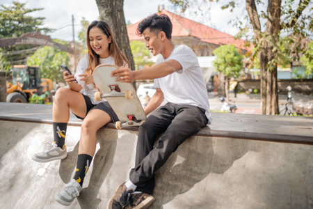 Two friends enjoy a fun moment at the skate park, immersed in their phones, laughing togetherの写真素材
