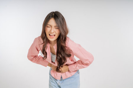 A young woman shows a grimace of pain while clutching her stomach in a studio settingの写真素材