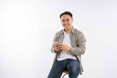 A cheerful young man is happily using his smartphone while comfortably seated on a chair