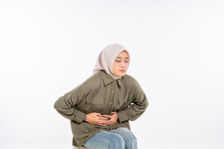 A woman in a hijab sits, holding her stomach, showing clear discomfort and painの写真素材