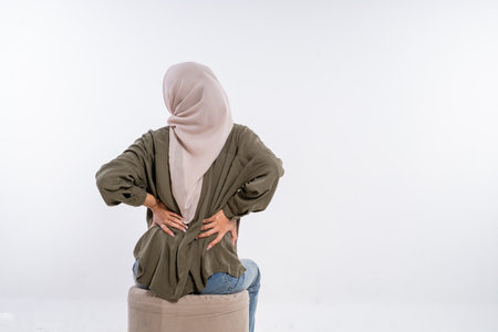 A woman in a headscarf sits on a stool, showing discomfort in her back and postureの写真素材