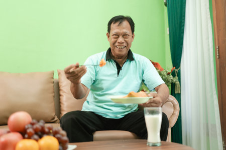An elderly man cheerfully enjoys a homemade meal at his cozy home, surrounded by fruitsの写真素材