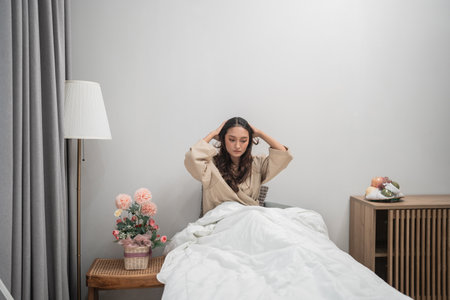A woman appears to be feeling stressed while sitting in a cozy and warm bedroom environmentの写真素材