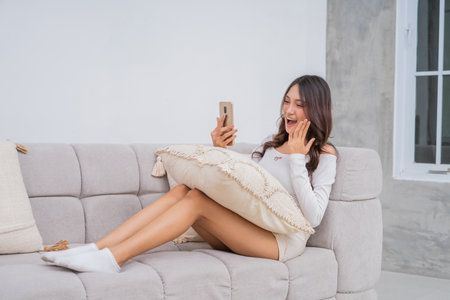 A young woman showcases her surprise while happily using her smartphone on a cozy couchの写真素材