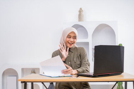 A woman happily smiles and waves while working on her laptop at home, embodying positivityの写真素材